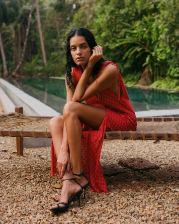 Model Lakshmi Menon wearing a red dress and black stiletto sandals seated on a woven daybed beside an outdoor pool.
