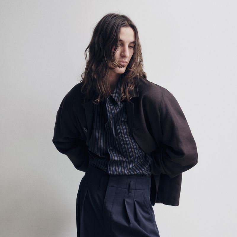 A young adult man with long brown hair models a striped button-down shirt, dark pants, and a blazer in a studio shot against a white wall.