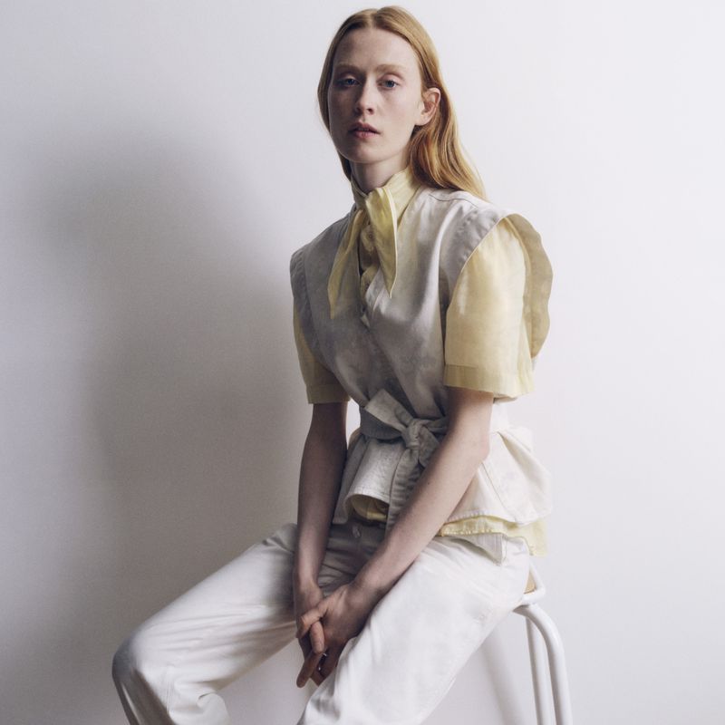 A red-haired adult female sits indoors on a white stool wearing a white and yellow linen shirt and white pants in a fashion setting.