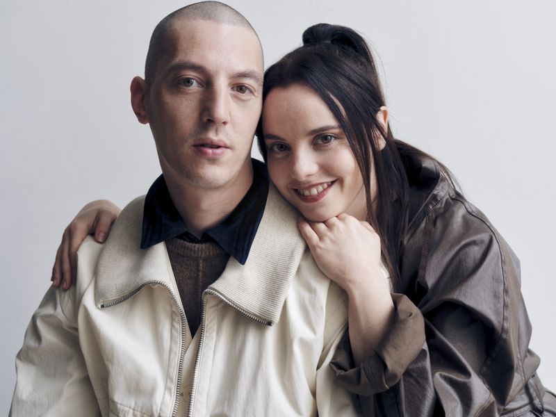 Portrait of Christina Ricci smiling while resting her chin on Mark Hampton's shoulder against a white studio background.