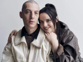 Portrait of Christina Ricci smiling while resting her chin on Mark Hampton's shoulder against a white studio background.
