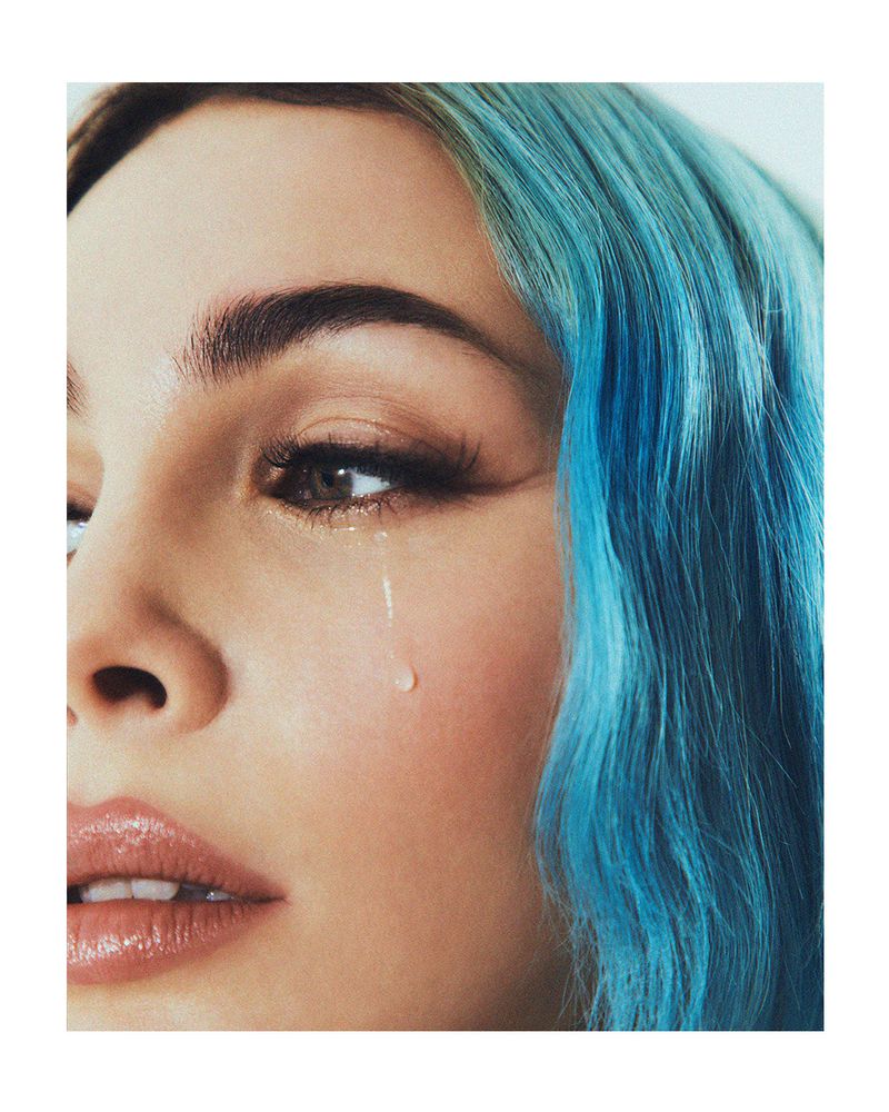 Close-up of a woman's face with blue hair and a tear running down her cheek, highlighting her eye with long lashes.
