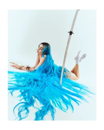Brunette woman with extra long blue hair lying down with a katana sword on a white background in a fashion or beauty editorial style photograph.