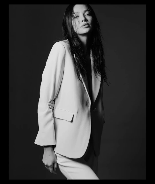 A black and white portrait of an Asian woman in a light-colored suit, posing against a dark background, conveying a classic and editorial style.