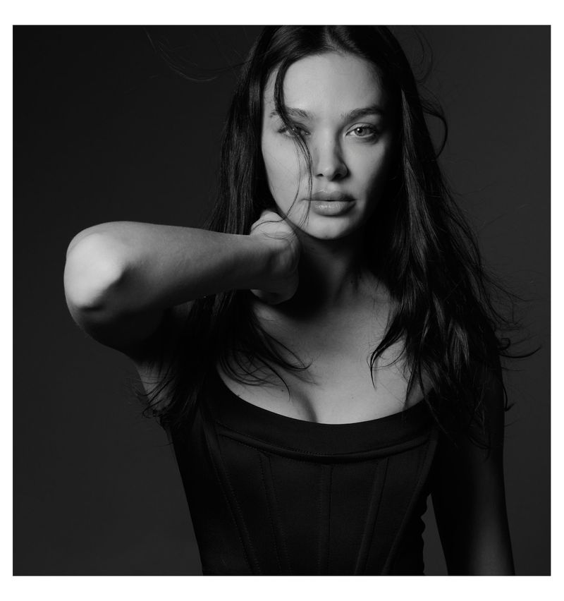 Black and white portrait of a young woman with dark hair looking at the camera, wearing a corset, and holding her fist to her neck.