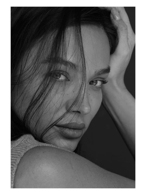Close-up black and white portrait of a woman looking at the camera with her hand behind her head and dark hair partially covering her face.