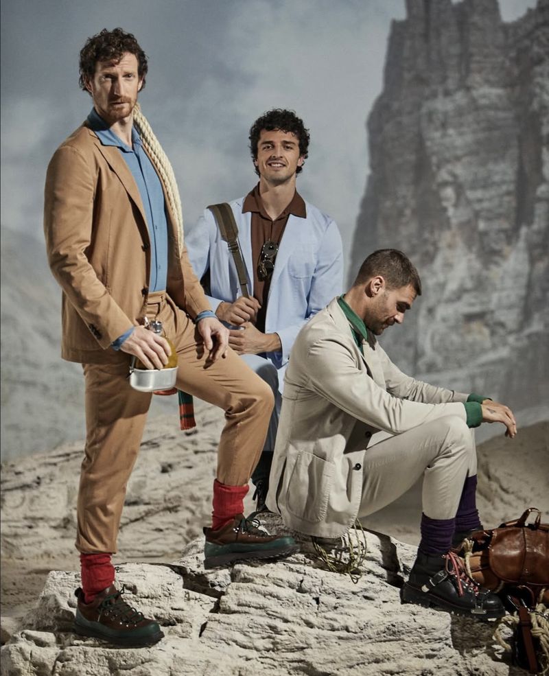 Three adult males pose in stylish outdoor apparel against a mountain backdrop, showcasing a blend of fashion and outdoor adventure.