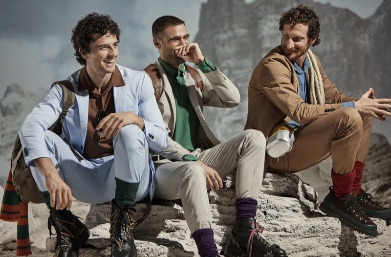 Three adult males in stylish suits pose outdoors on rocks, with a blurred mountain background, showcasing fashion and outdoor lifestyle.