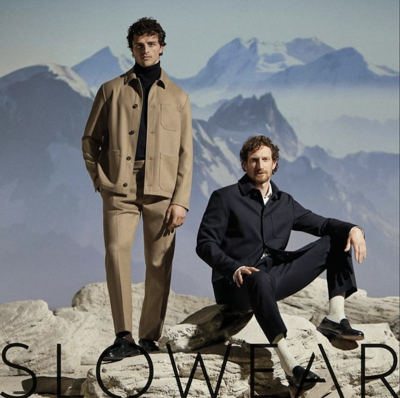 Two adult males pose in studio shot for fashion wear, one standing in a tan suit, the other sitting on rocks in a blue jacket.