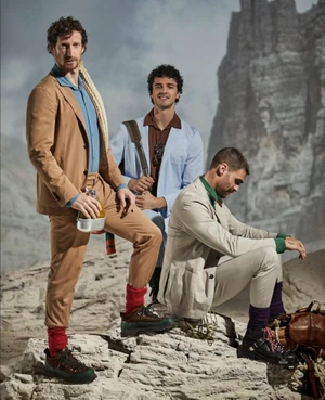 Three men wearing tailored suits and hiking boots pose on a rocky outcrop against a mountain background.