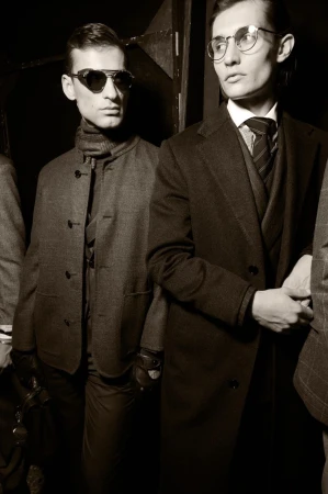 Three men in stylish vintage-toned attire including coats, suits, glasses, and leather gloves pose indoors in a vertical medium shot.