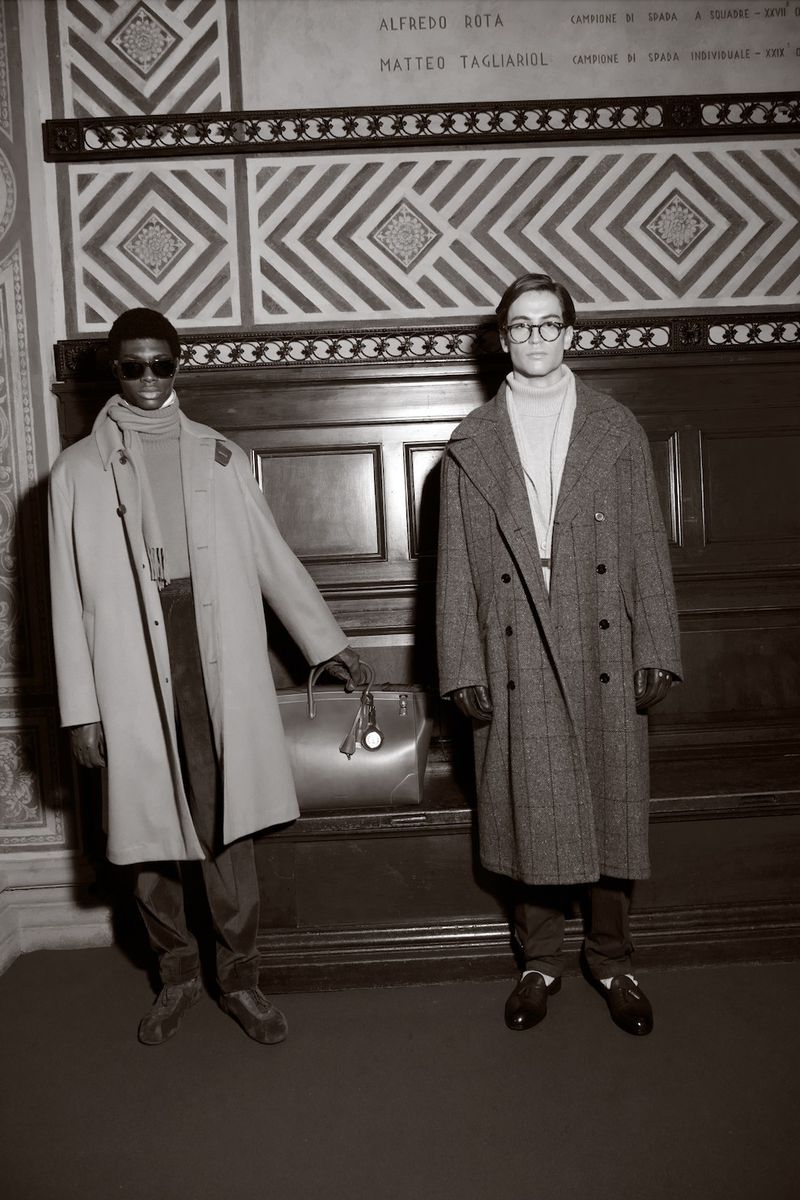 Two men in overcoats pose indoors with a leather bag, captured in a subdued, desaturated photograph highlighting fashion and style.