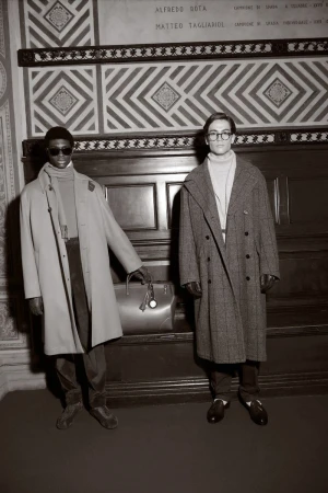 Two men in overcoats pose indoors with a leather bag, captured in a subdued, desaturated photograph highlighting fashion and style.