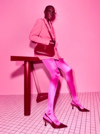 A fashion photo features a black woman with glasses, wearing a pink blazer and pink patterned tights, posing next to a table against a pink backdrop.