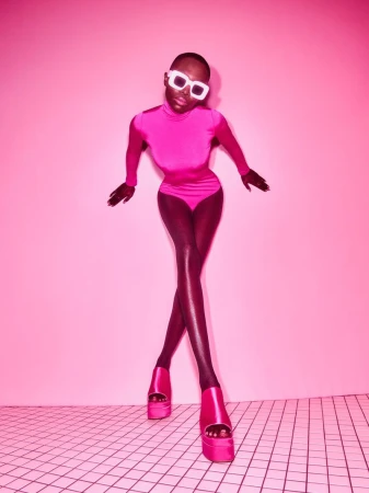 Amina Seck wearing a bright pink turtleneck bodysuit, white sunglasses, and pink platform heels against a pink background on tiled floor.
