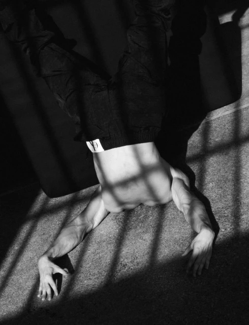 Black and white studio shot of a shirtless adult male performing a handstand against a wall with shadows cast across his body.