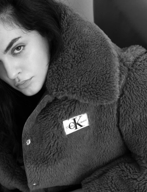 Monochrome portrait of a young adult woman wearing a teddy coat with a visible Calvin Klein logo. Eye-level shot in diffused light.