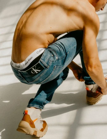 A shirtless man in blue jeans and boots is kneeling in a photo studio in an editorial fashion shot featuring a Calvin Klein waistband.