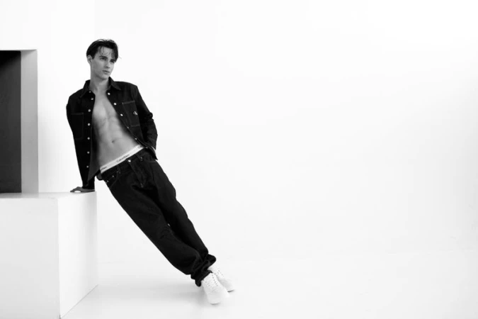 Model Iasonas Laios leaning against a white plinth wearing an open black jacket and jeans for Calvin Klein.