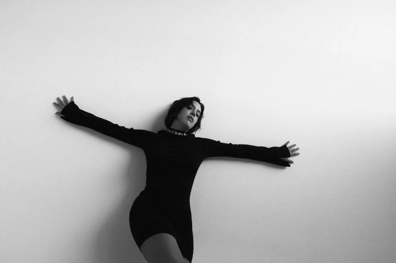 Monochrome studio shot of a young woman with short dark hair in a little black dress, arms outstretched touching a white wall.