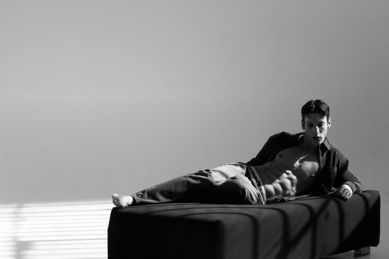 Black and white photo of a muscular adult male lying on a black upholstered surface, wearing an unbuttoned shirt and pants.