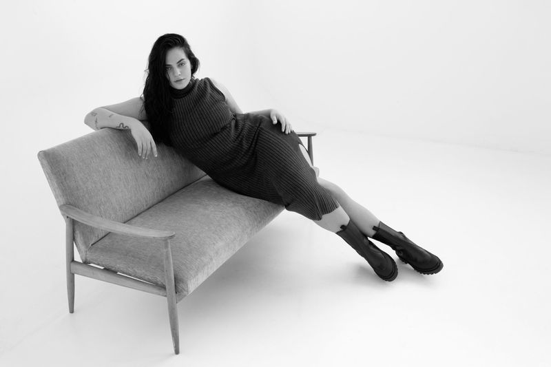 Model with long dark hair reclining on a mid-century sofa wearing a ribbed dress and boots in a studio.