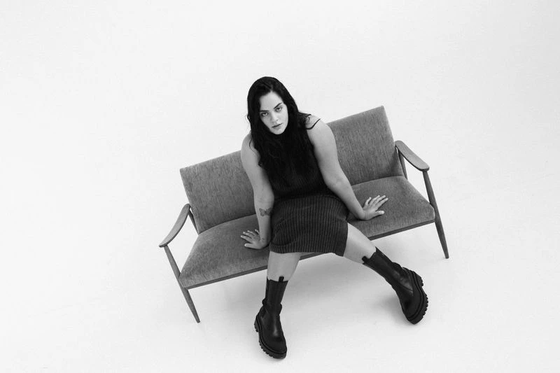 A stylish adult female sits on a grey two-seater sofa against a bright white backdrop. She wears a dark top, knit skirt, and boots.