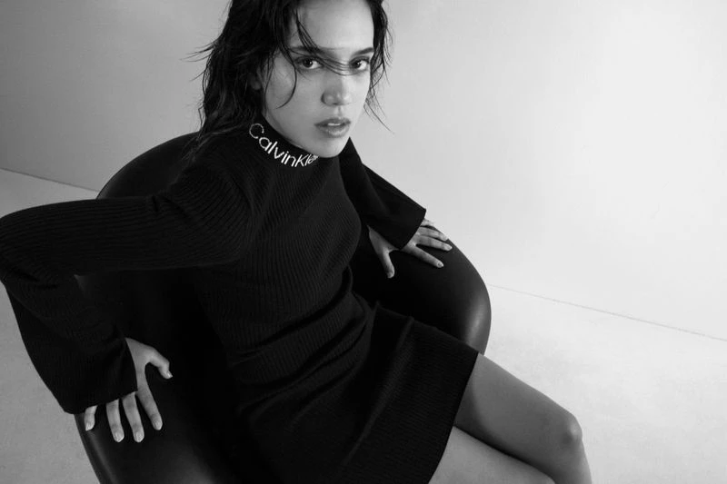 A woman with wet hair and a black turtleneck dress with a Calvin Klein logo poses in a black chair against a light grey background.