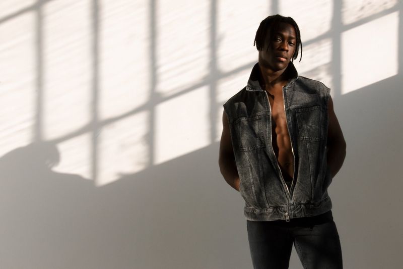 Portrait of Black adult male in denim vest standing in front of a wall with window shadow patterns, fashionable casual pose.