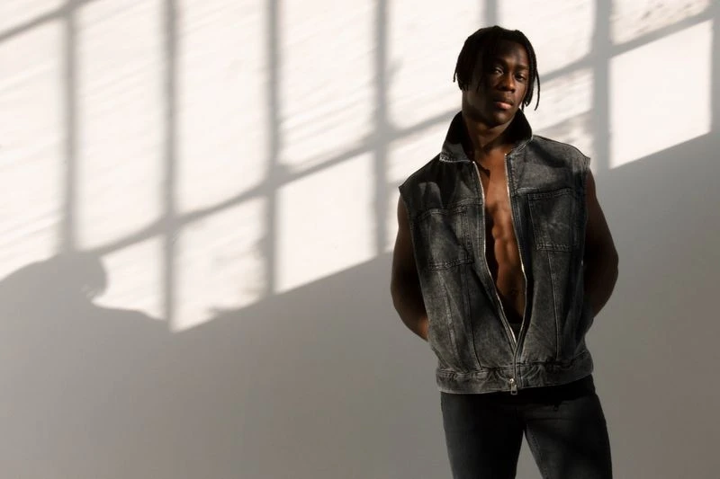 Portrait of Black adult male in denim vest standing in front of a wall with window shadow patterns, fashionable casual pose.