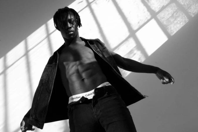 Black and white portrait of a shirtless adult male wearing a denim vest, jeans, and Calvin Klein underwear in a studio setting.