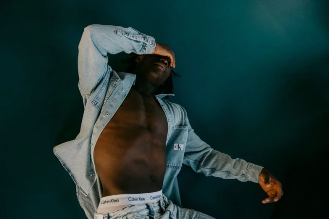 An African American adult male poses in a studio, wearing a light denim jacket, and jeans with the 'Calvin Klein' logo visible.