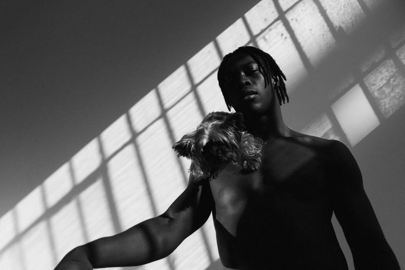 Black and white portrait of a shirtless Black man with braids holding a small dog on his shoulder under window shadows.