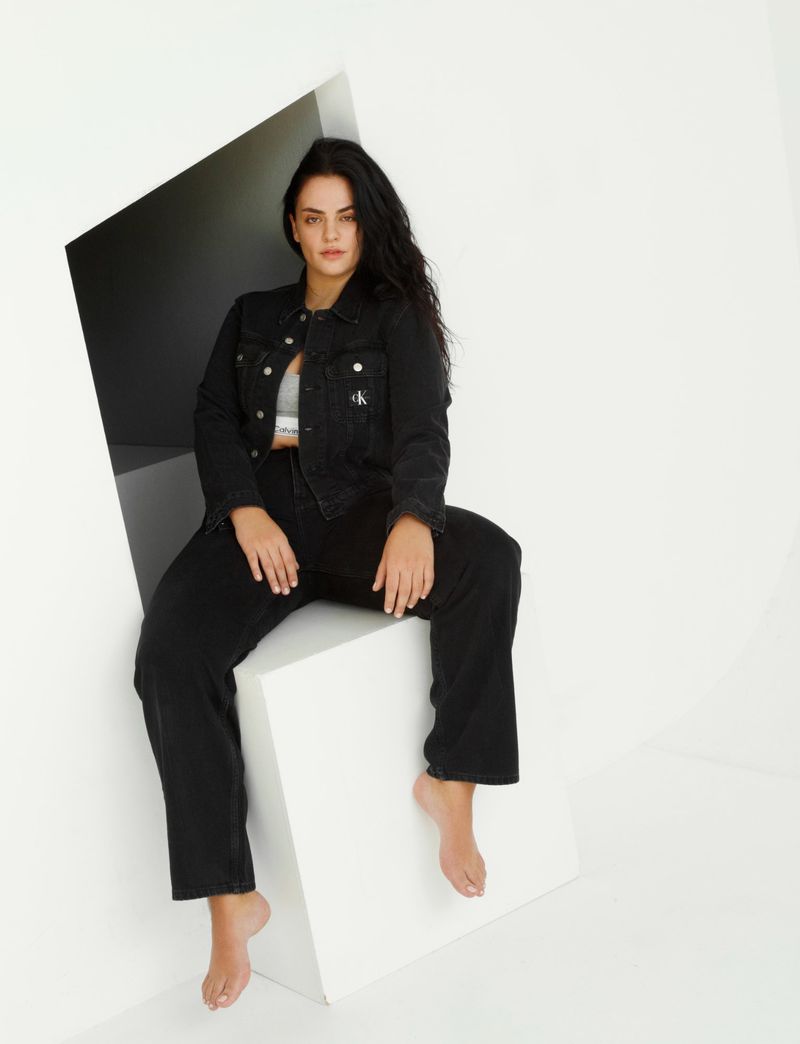 A woman in a black denim outfit sits on a white cube in a minimalist studio. She wears a grey top and is barefoot, with long dark hair.