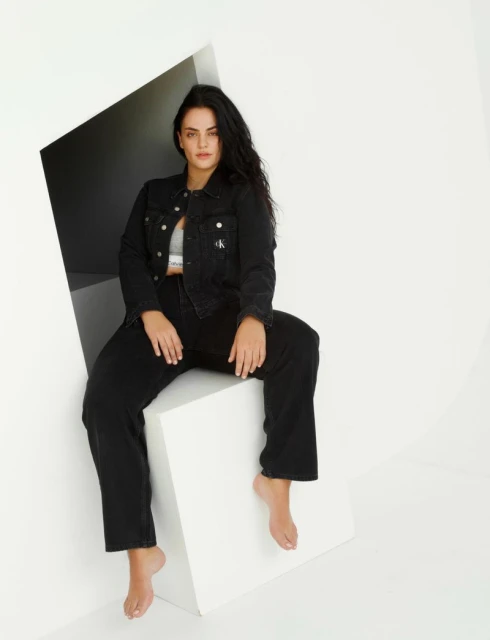 A woman in a black denim outfit sits on a white cube in a minimalist studio. She wears a grey top and is barefoot, with long dark hair.