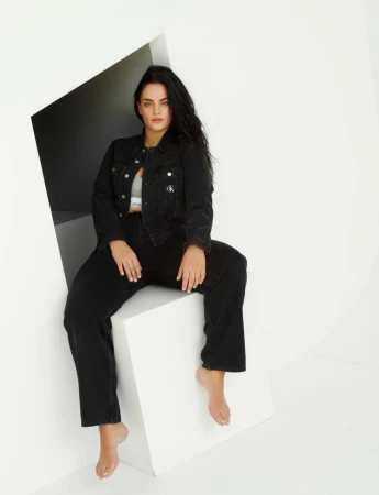A woman in a black denim outfit sits on a white cube in a minimalist studio. She wears a grey top and is barefoot, with long dark hair.