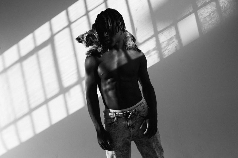 A shirtless man with dreadlocks and jeans poses indoors with a small dog on his shoulders, lit by natural light casting shadows on the wall.