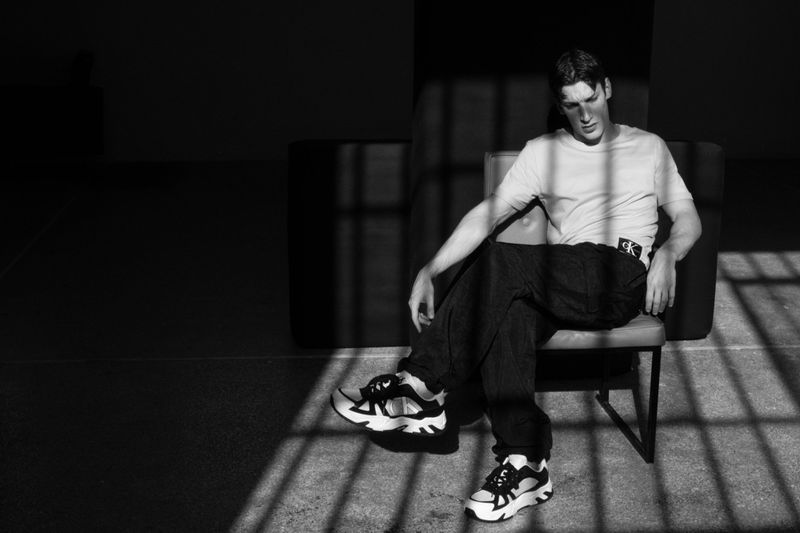 Monochrome shot of a man in a t-shirt and dark pants sitting on a chair, with light streaming through window bars in the background.