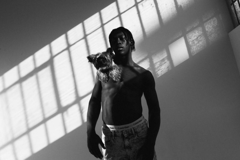 Monochromatic portrait of a bare-chested African American man holding a small dog with window shadow patterns in a bright indoor setting.