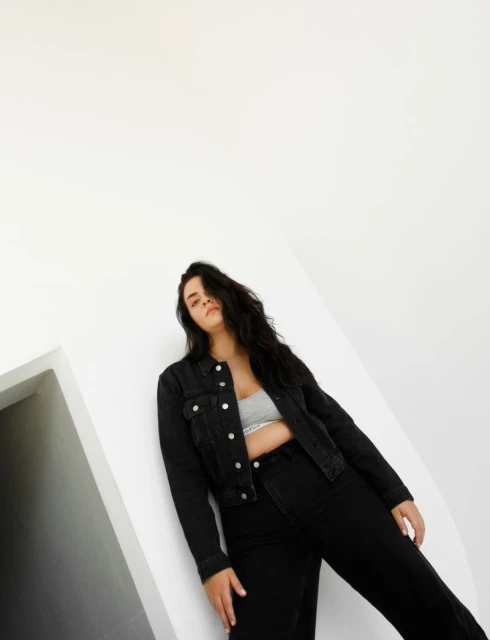 A woman with long, dark hair poses in a black denim jacket and pants against a white background, styled with a modern fashion aesthetic.