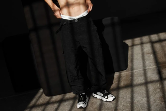 A person poses indoors in black pants and white athletic shoes. Calvin Klein branded underwear is visible above the pants.