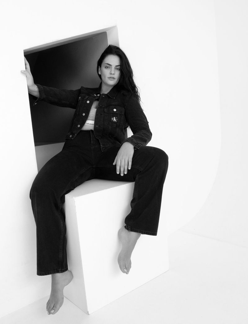 Model wearing a black denim jacket and matching jeans seated barefoot on a white geometric box structure in a studio.