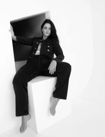 Model wearing a black denim jacket and matching jeans seated barefoot on a white geometric box structure in a studio.