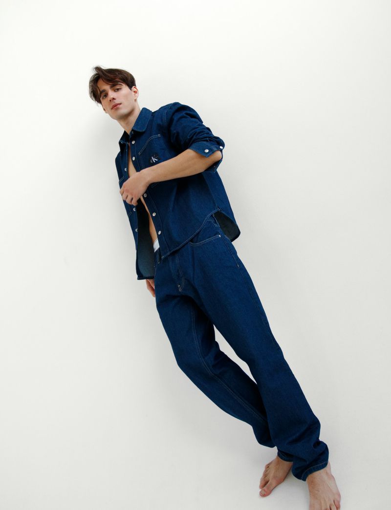 Barefoot man in unbuttoned dark blue denim shirt and matching pants poses against a white wall looking at the camera.
