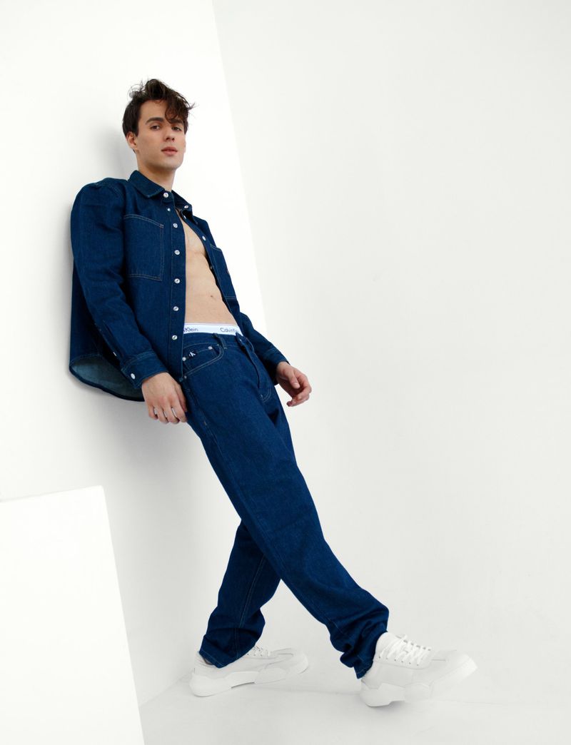 A young adult male poses in a denim shirt, jeans, and white sneakers against a bright white wall in a studio setting with natural lighting.