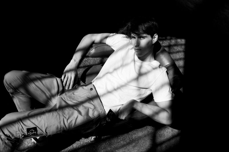 Monochrome portrait of a young adult man lounging on a dark leather couch, wearing a white t-shirt and pants, with window shadows.