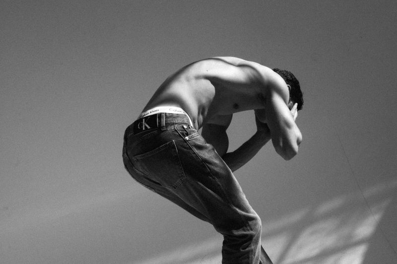 Monochrome portrait of athletic man with bare chest, wearing jeans and a Calvin Klein waistband, in a crouched pose against gradient backdrop.