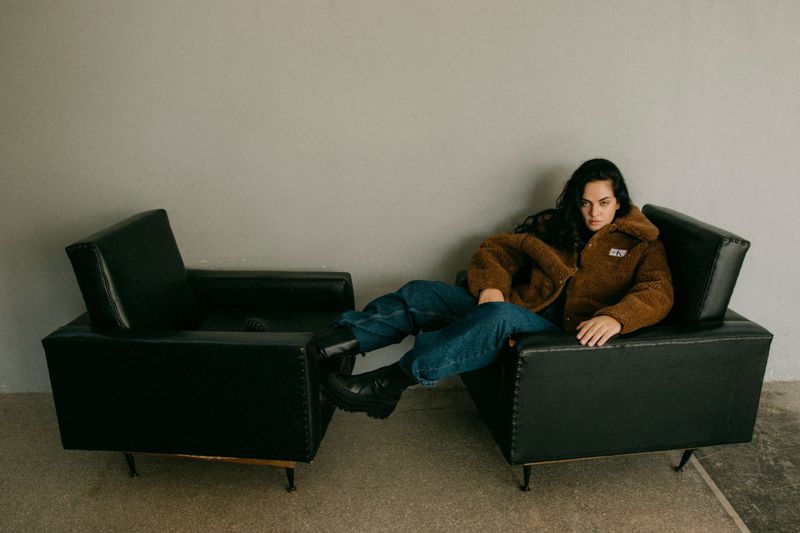 Woman in a brown jacket and blue jeans sitting in a black armchair in a simple indoor setting, creating a casual and relaxed atmosphere.