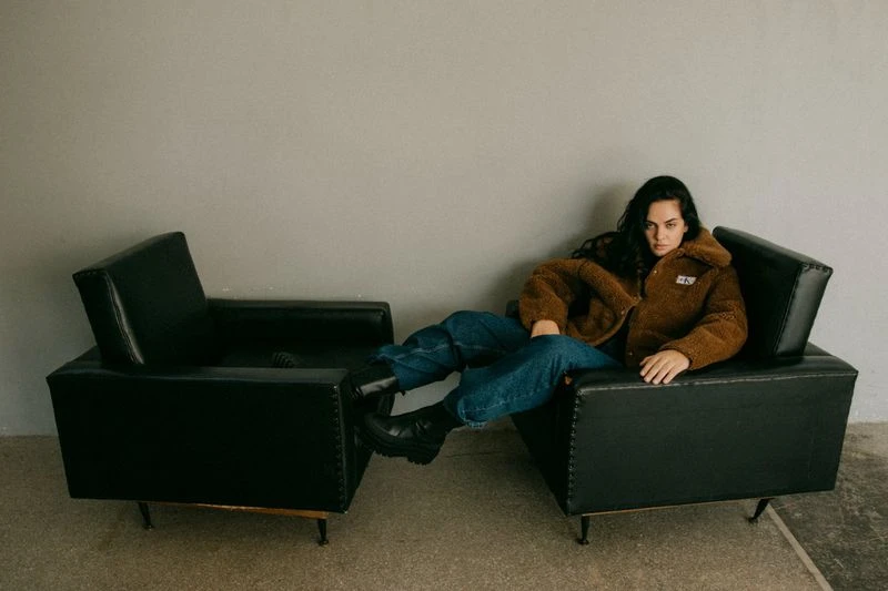 Woman in a brown jacket and blue jeans sitting in a black armchair in a simple indoor setting, creating a casual and relaxed atmosphere.