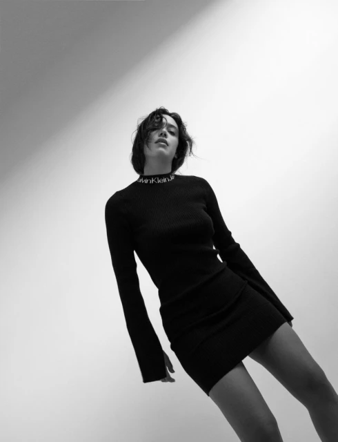 Black and white portrait shot of a young woman in a long-sleeve black mini dress against a light backdrop, posing at a low angle.